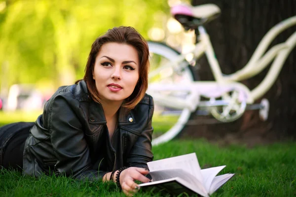 Mujer joven con libro de lectura de bicicletas en el parque de la ciudad en la hierba — Foto de Stock