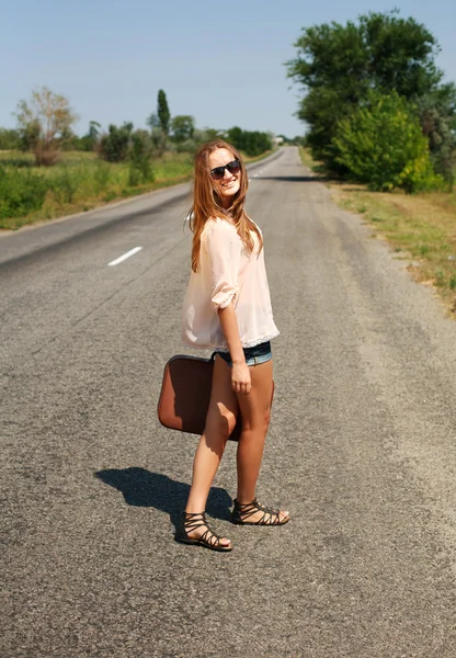 Jonge vrouw met koffer liften op weg op platteland — Stockfoto