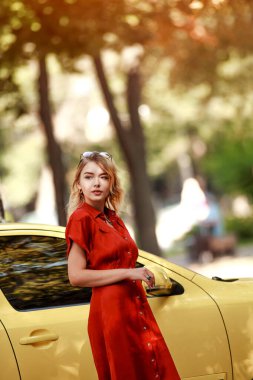 Şehir parkında sarı bir arabanın yanında kırmızı elbiseli, çekici ve hayalperest bir iş kadını.