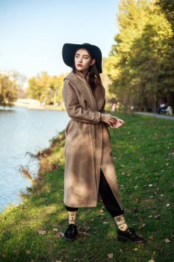 Uzun bej ceketli, siyah şapkalı şık bir bayan yeşil bir parkta nehir kenarında yürüyor.