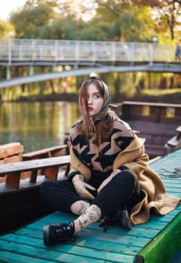 Uzun paltolu zarif genç bayan yeşil parktaki teknenin yanındaki ahşap iskelede oturuyor.