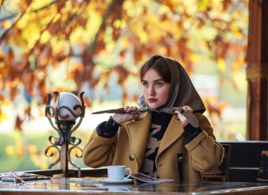 Restoranın yaz terasındaki masasında bej ceketli ve türbanlı genç bir iş kadını. Yanında bir fincan kahve ve bir dergi, şık bir bayanın portresi.