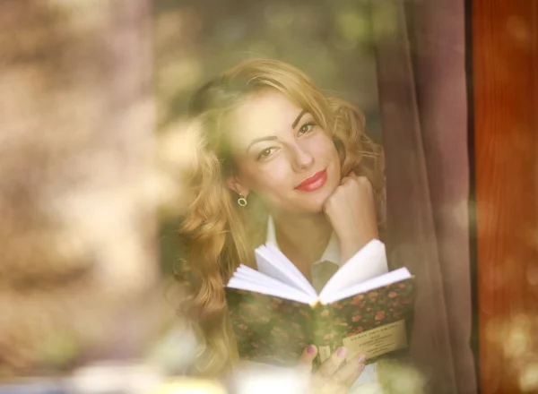 Mulher sorridente com livro em casa, vista através de vidro de janela — Fotografia de Stock