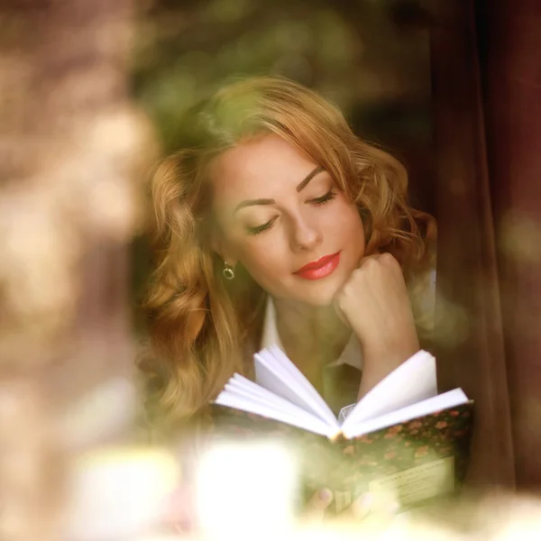 Piękna dziewczyna czytając książkę pomieszczeniu, widok przez okno — Zdjęcie stockowe