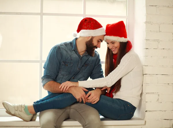 Glückliches junges Paar in roten Weihnachtsmützen am Fenster sitzend — Stockfoto