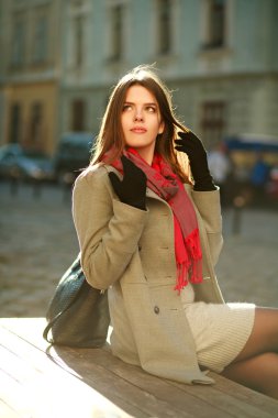 Güneş ışığında caddenin üzerinde oturan paltolu güzel kadın