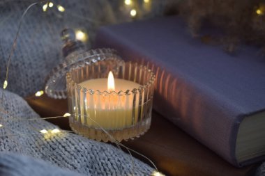 Candle with closed book and garland on background of knitted gray blanket with warm lights creates an evening homey atmosphere.