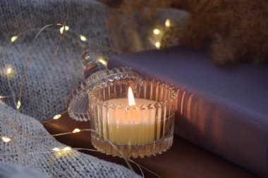 Candle in glass jar on warm blanket with book. Cozy evening. Winter or autumn atmosphere.