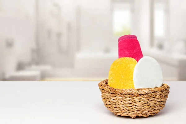 Basket with three colorful terry towels or cosmetic for body care on a white table over blurred bath background with copy space. For your product display montage.