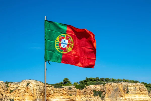 Selective focus on waving flag of portugal in front of typical yellow rock formation of algarve and blue sky. National portugal flag with space.