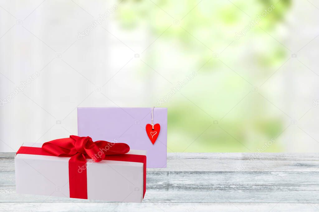 Valentines day background. Gift box with red ribbon and a letter with a red heart on a wooden tabletop over a blurred bright window background. Space for design.