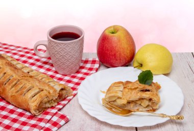 Elmalı dürüm. Bir rulo ev yapımı taze elmalı turta, bir fincan çay, tabakta iştah açıcı bir dilim elmalı turta ve arka planda taze elmalar. Kırsal kahvaltı konsepti.