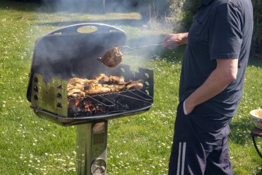 Kömür lokantasında bir adam biftek, ızgara sosis ve tavuk kanadı pişiriyor ve barbekü maşası kullanıyor. Güneşli bir bahar gününde yeşil çayırda leziz bir barbekü hazırlığı. Barbekü..