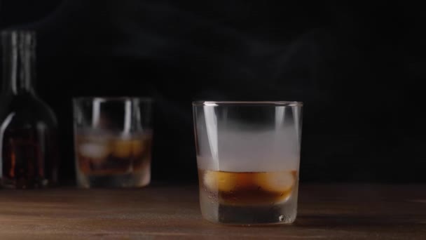 Glas med whisky och is i rök på träbord. — Stockvideo