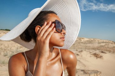 Koyu tenli, güneş gözlüklü bir kadın yazın çölde kumların üzerinde yürüyor.