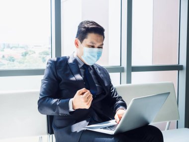 Koruyucu yüz maskesi takan Asyalı işadamı, modern ofis binasında büyük cam pencerenin yanında otururken dizüstü bilgisayarının monitörünü görünce mutlu ve neşeli bir şekilde elini kaldırdı..