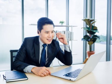 Takım elbiseli Asyalı işadamı ofiste dizüstü bilgisayar kullanıyor. Kulaklıklı adam, mikrofon görünümlü bilgisayar ekranlı konferans çağrısı, online toplantı ve müşteri destek operatörü konsepti.