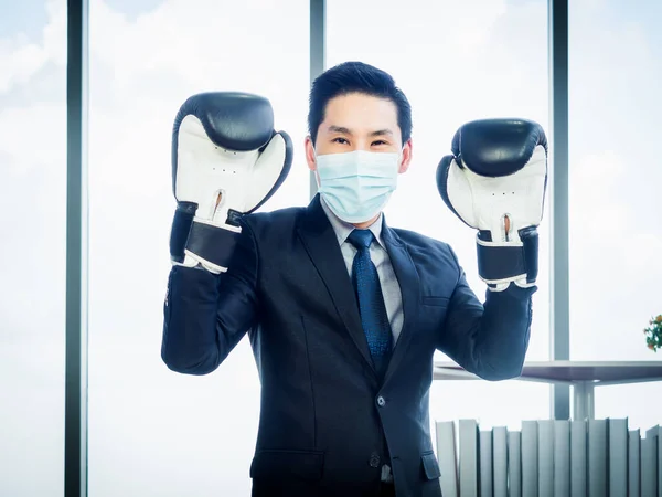 Asian Businessman wearing suit and protective face mask and hand wearing boxing gloves in office. Man wears mitten, posture in boxing ready to fight the problem concept.