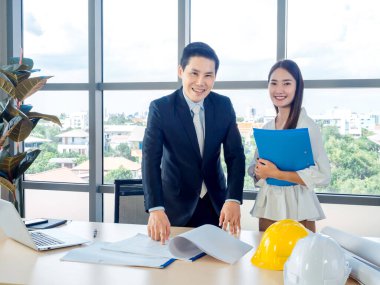 Asyalı erkek mimar ya da mühendis takım elbiseli ve genç bayan sekreter planda dizüstü bilgisayarlı ve masasında beyaz ve sarı başlıklı büyük cam pencereli ofislerde tartışırlar..