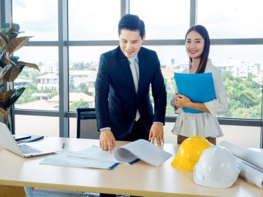 Asyalı erkek mimar ya da mühendis takım elbiseli ve genç bayan sekreter planda dizüstü bilgisayarlı ve masasında beyaz ve sarı başlıklı büyük cam pencereli ofislerde tartışırlar..