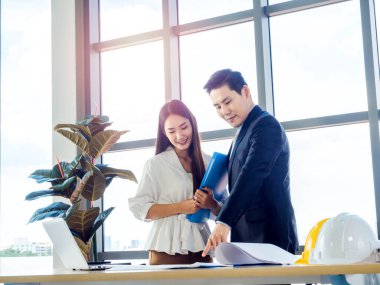 Asyalı erkek mimar ya da mühendis takım elbiseli ve genç bayan sekreter planda dizüstü bilgisayarlı ve masasında beyaz ve sarı başlıklı büyük cam pencereli ofislerde tartışırlar..