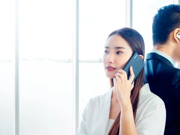 Business Asian couple talking to mobile phone. Young beautiful confident woman calling with cell phone and standing back with businessman in suit on huge glass window background with copy space.