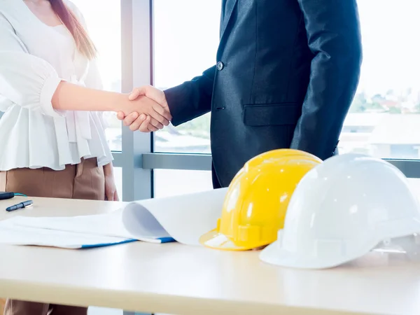 Handshake of collaboration concept. Businessman in suit, engineering or architect and woman shaking hands on blueprint and yellow and white safety hard hat on desk on glass window background.