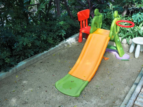 Old empty orange and green color playground slide for kid on sand near the garden.