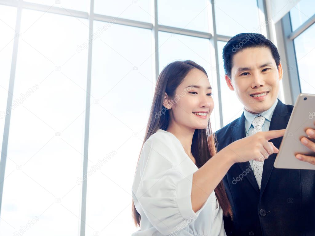 Business couple working together. Asian businessman in suit and smiling young woman pointing at digital tablet while both watching at screen on glass window background with copy space.