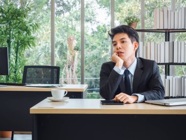 Takım elbiseli, yorgun, stresli ve üzgün bir Asyalı genç işadamı ofisinde dalgın dalgın oturuyor. Hüsrana uğramış işçi zihinsel sağlık sorunları ve tükenme sendromu kavramı.