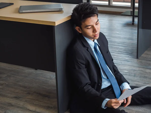 Young Asian businessman receiving bad news with dismissal letter, tired, stressed and sad sitting with absent-minded on the ground in office. Frustrated worker mental health problems.