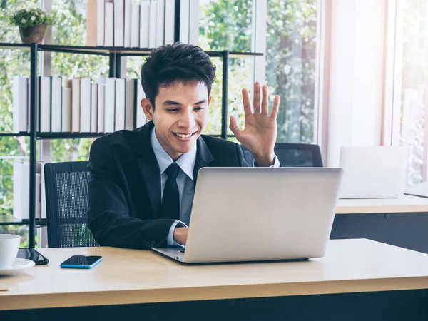 Young smiling Asian businessman in suit greeting someone, wave to camera on laptop computer have video conversation online while working on desk in office near huge glass window with sunlight.