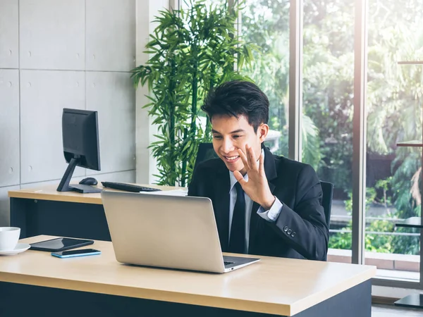 Young smiling Asian businessman in suit greeting someone, wave to camera on laptop computer have video conversation online while working on desk in office near huge glass window with sunlight.