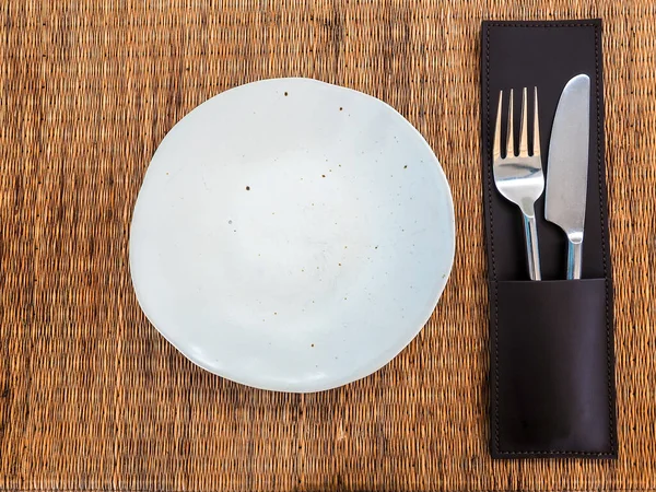 Ceramic plate. Empty white freeform shaped porcelain dish with stainless knife and fork in leather case on mat, top view.