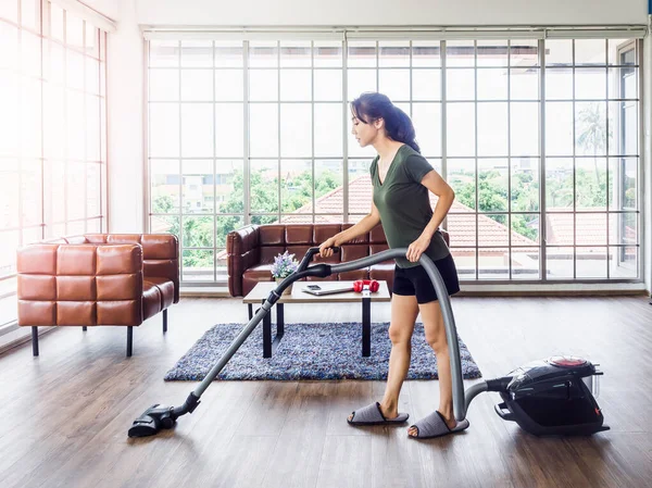 Young beautiful Asian woman, housewife wearing casual clothes using a vacuum cleaner to clean floor and carpet in living room near brown leather sofa and huge glass window inside house on sunny day.