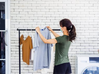 Genç, güzel Asyalı bir kadın, koruyucu maske takan koruyucu bir ev hanımı çamaşır makinesinin yanında çamaşır makinesinin yanında çamaşır ipiyle asılmış kuru bir gömlek asıyor..