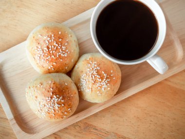 Tahta tepside beyaz susam tohumlu ev yapımı taze hamburger çörekleri ve kahvaltı için ahşap masada sıcak kahve, üst manzara. Fırından yeni çıkmış üç lezzetli sandviç ekmeği..