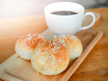 Kahvaltı için ahşap masada sıcak kahve ile servis edilen küçük tahta tepside beyaz susam tohumlu ev yapımı hamburgerler. Fırından yeni çıkmış üç lezzetli sandviç ekmeği..