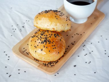Küçük tahta tepside siyah susamlı taze ev yapımı hamburger çörekleri ve beyaz masa örtüsü üzerinde sıcak kahve servisi. Fırından yeni çıkmış iki lezzetli sandviçli çörek..