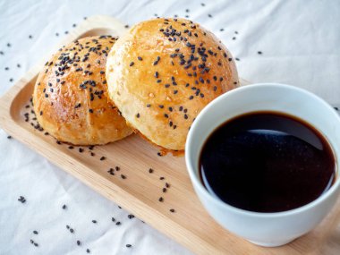 Küçük tahta tepside siyah susamlı taze ev yapımı hamburger çörekleri ve beyaz masa örtüsü üzerinde sıcak kahve servisi. Fırından yeni çıkmış iki lezzetli sandviçli çörek..