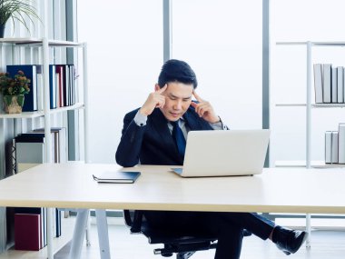 Takım elbiseli yakışıklı Asyalı işadamı, ekranı seyredip parmağıyla kafasını işaret ederken kitaplığın ve cam pencerenin yanındaki masada dizüstü bilgisayarla çalışırken düşünüyor..