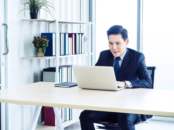 Handsome Asian businessman in suit working with laptop computer on desk inside the white office container house near book shelf and glass window. Man manager using laptop in well-lit workplace.