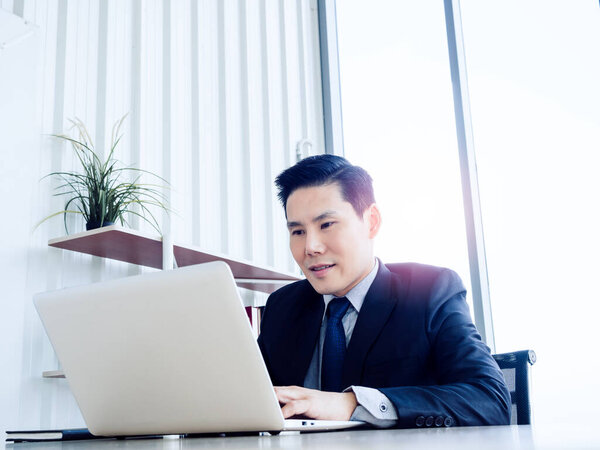 Handsome Asian businessman in suit working with laptop computer on desk inside the white office container house near glass window. Man manager using laptop in well-lit workplace.