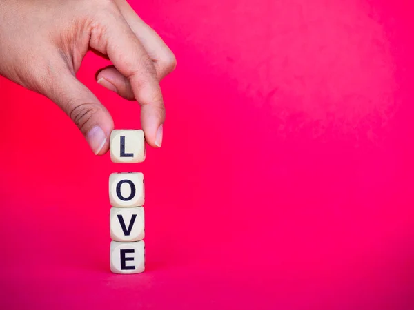 Love word written on wood block on pink red background with copy space, minimal style. Hand holding L to put on Love, text is written in black letters on wooden cubes.