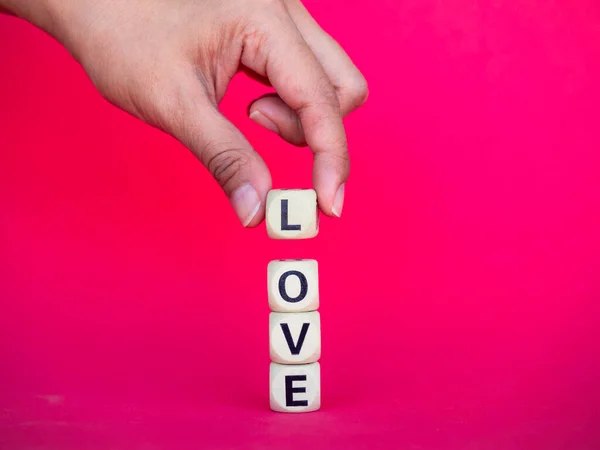 Love word written on wood block on pink red background, minimal style. Hand holding L to put on Love, text is written in black letters on wooden cubes.