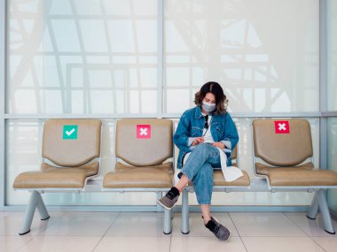 Güzel Asyalı kadın ameliyat maskesi ve kot ceket giyiyor. Akıllı telefon kullanıyor ve bekleme sandalyelerinde oturuyor. Sosyal uzaklık işareti, yeşil onay işareti ve havaalanında fotokopi alanı olan kırmızı haç..