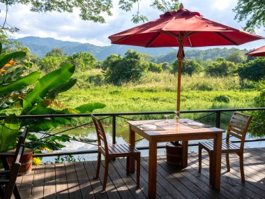 Yemek takımı ve şarap kadehiyle boş ahşap yemek masası. Masa, sandalyeler ve rıhtımdaki ahşap terasta kırmızı şemsiye. Dağ ve güzel yeşil doğa manzaralı..
