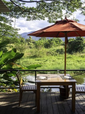 Yemek takımı ve şarap kadehiyle boş ahşap yemek masası. Masa ve sandalyeler ve rıhtımdaki ahşap terastaki kırmızı şemsiye dağ ve güzel yeşil doğa manzaralı, dikey stil..