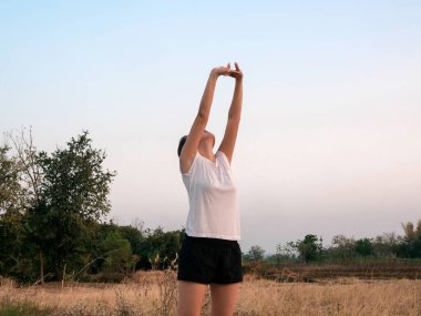 Genç Asyalı kadın çimen tarlasının yanında kollarını mavi gökyüzüne uzatıyor. Kadın egzersizden önce dışarıda ısınıyor. Fitness ve sağlıklı yaşam tarzı konsepti.