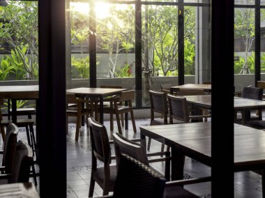 Cam odada boş ahşap yemek masaları sabah güneş ışığıyla kahvaltıya gelen misafirler için hazırlanıyor. Ahşap yemek masası ve dışarıdaki yeşil bahçenin yanında sandalyeleri olan kapalı bir restoran..